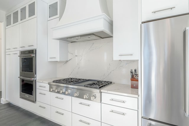 kitchen featuring decorative backsplash, custom range hood, glass insert cabinets, light stone counters, and stainless steel appliances