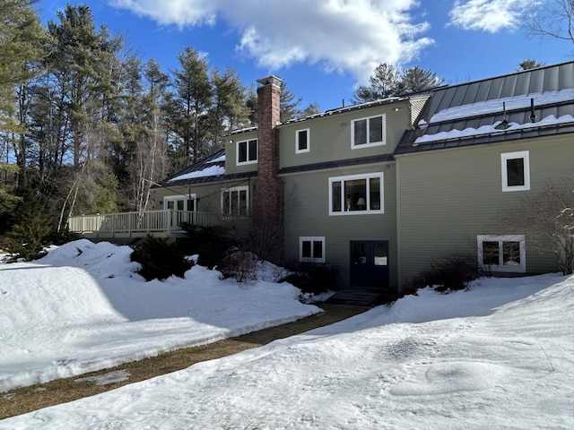 snow covered rear of property featuring a standing seam roof, a deck, a chimney, and metal roof