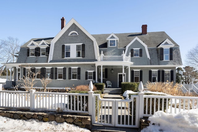 shingle-style home with a gambrel roof, a fenced front yard, a chimney, roof with shingles, and a porch