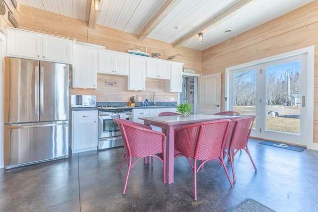 interior space with wooden walls, concrete floors, beam ceiling, white cabinets, and appliances with stainless steel finishes