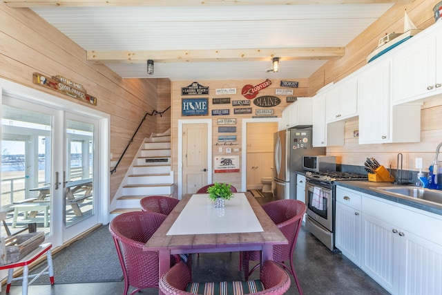 kitchen featuring wooden walls, beam ceiling, white cabinets, stainless steel appliances, and a sink