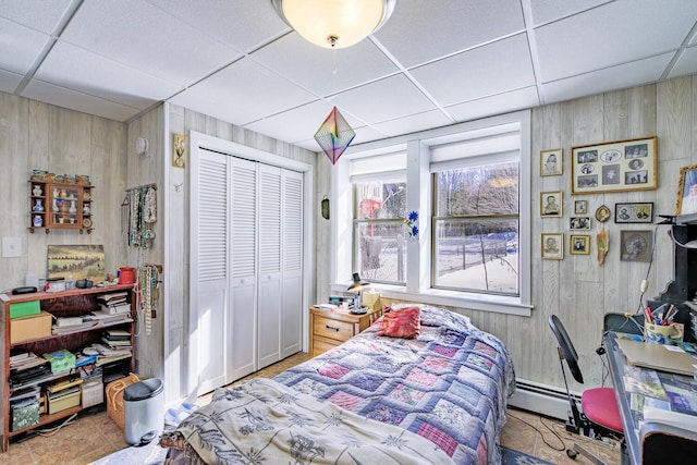 bedroom with a closet, a drop ceiling, wood walls, and baseboard heating