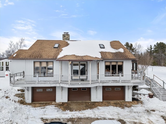 snow covered back of property with a shingled roof, a chimney, an attached garage, and stairs