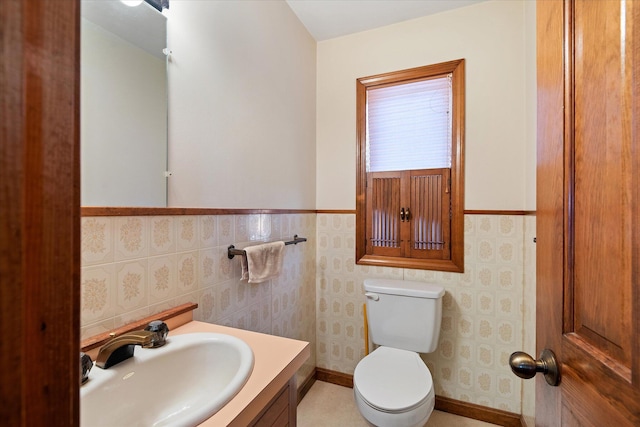 half bathroom featuring wallpapered walls, baseboards, wainscoting, toilet, and vanity