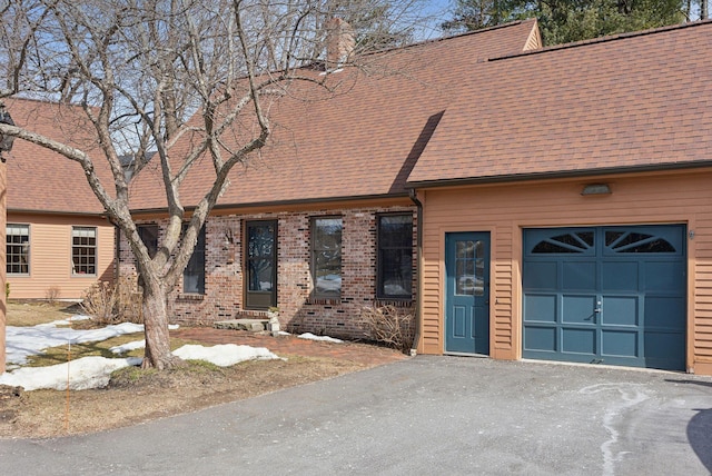 view of front of house featuring aphalt driveway, roof with shingles, a garage, brick siding, and a chimney