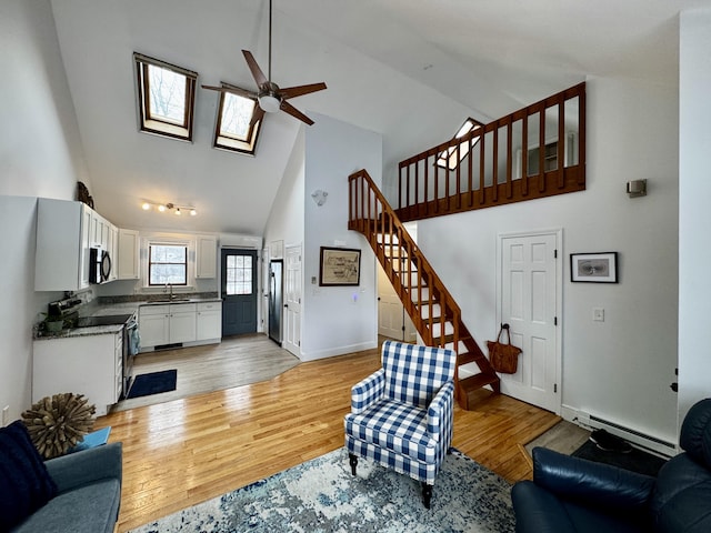 living area with light wood-style flooring, ceiling fan, stairway, baseboard heating, and high vaulted ceiling