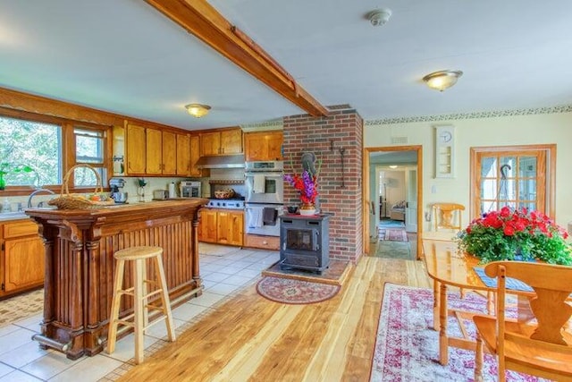 kitchen featuring brown cabinetry, stainless steel double oven, a wood stove, beamed ceiling, and under cabinet range hood