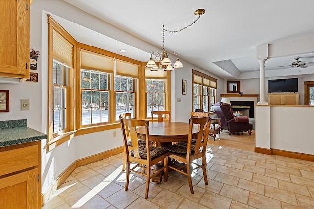 dining area with baseboards, a glass covered fireplace, stone finish floor, ornate columns, and ceiling fan with notable chandelier