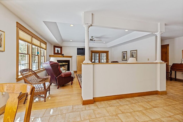 interior space featuring a ceiling fan, a glass covered fireplace, a tray ceiling, and ornate columns