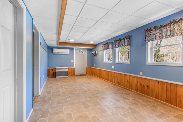 empty room with wooden walls, stone finish flooring, a wall unit AC, wainscoting, and a paneled ceiling