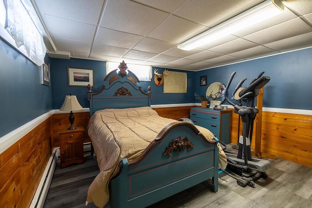 bedroom with a drop ceiling, wainscoting, a baseboard radiator, and wooden walls