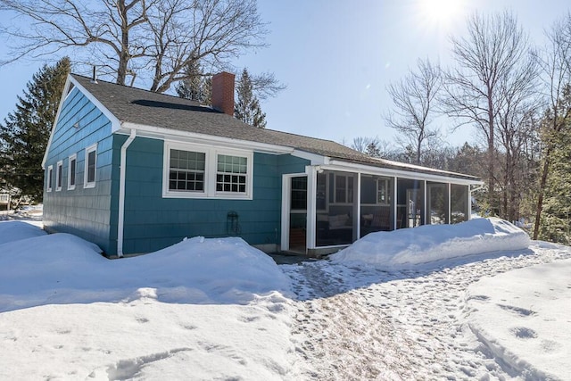 snow covered back of property featuring a sunroom, roof with shingles, and a chimney