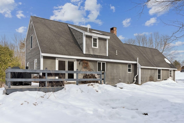 snow covered house with a deck, a shingled roof, and a chimney