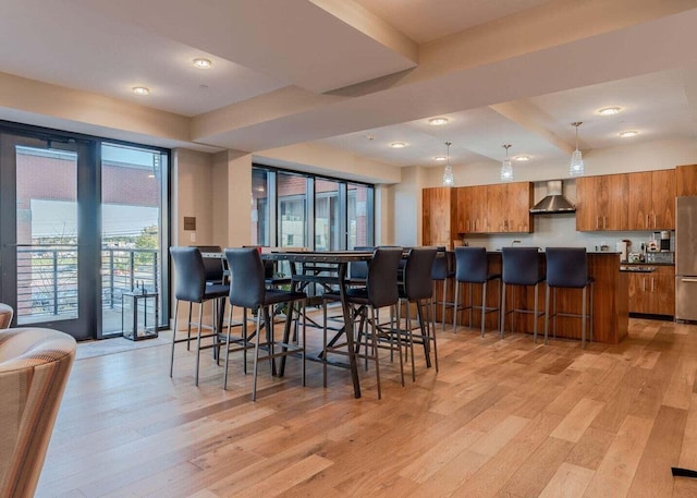 kitchen with light wood finished floors, brown cabinetry, wall chimney exhaust hood, a breakfast bar area, and freestanding refrigerator