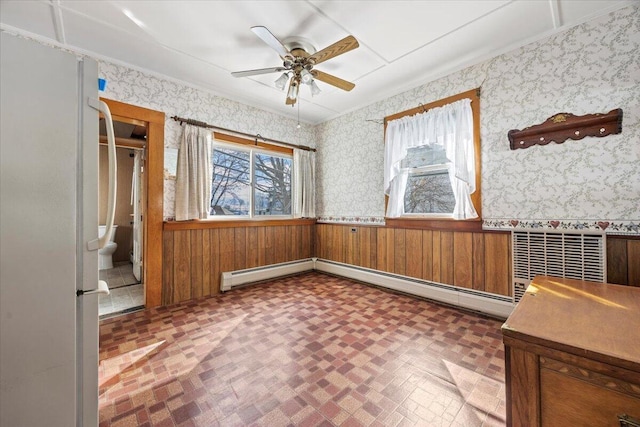 spare room featuring a baseboard heating unit, wainscoting, ceiling fan, and wallpapered walls