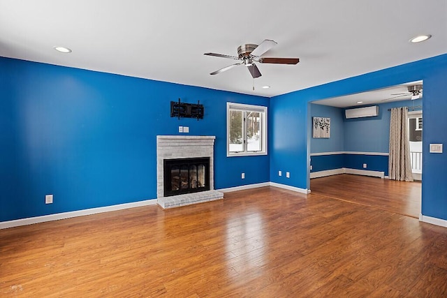 unfurnished living room featuring a fireplace, a wall mounted AC, a ceiling fan, wood finished floors, and baseboards