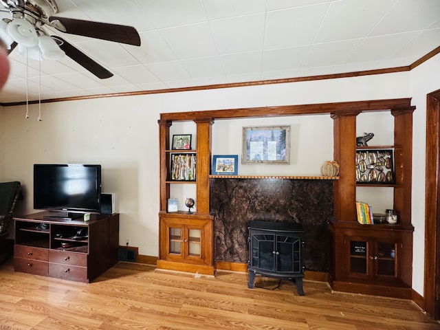 living room featuring wood finished floors, baseboards, a wood stove, ceiling fan, and crown molding