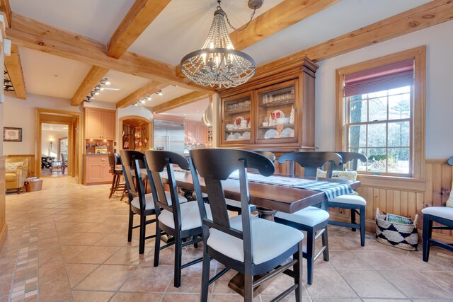 dining space with beam ceiling, light tile patterned flooring, a chandelier, and wainscoting