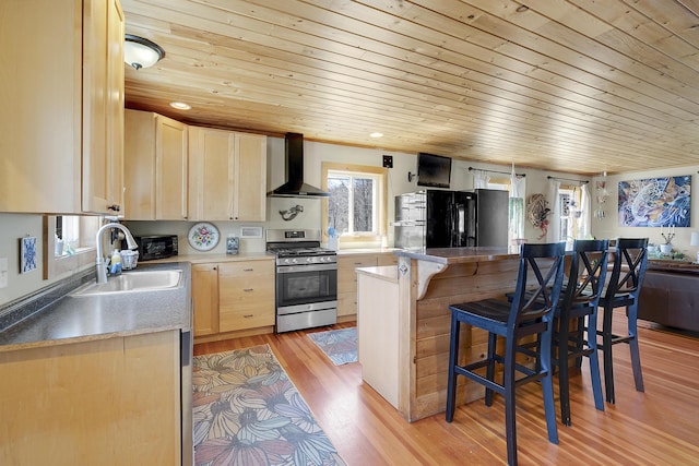 kitchen with freestanding refrigerator, stainless steel gas stove, light brown cabinets, wall chimney exhaust hood, and a sink