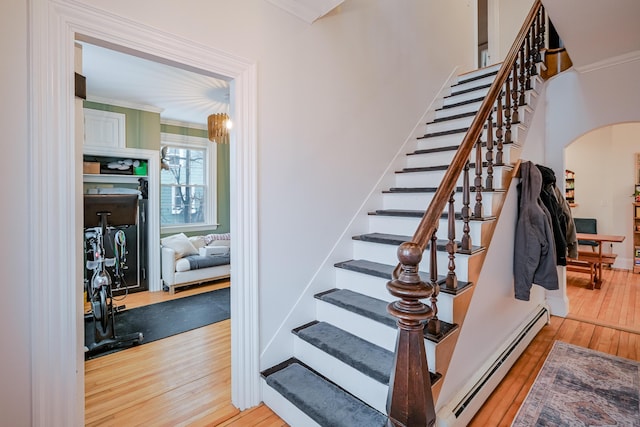 stairs with wood finished floors, arched walkways, baseboard heating, and ornamental molding