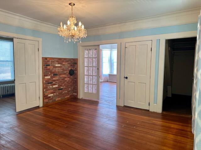 unfurnished dining area featuring wood finished floors, radiator, an inviting chandelier, brick wall, and crown molding