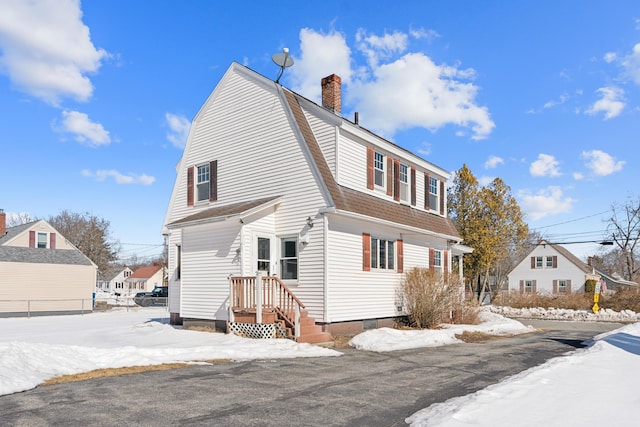 snow covered property with a gambrel roof, roof with shingles, a chimney, and fence