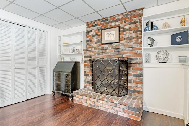 living room featuring a brick fireplace, built in features, wood finished floors, and a drop ceiling