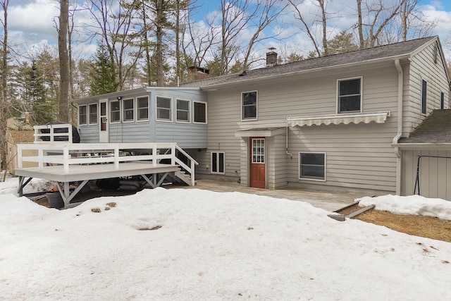 snow covered rear of property featuring a wooden deck, a chimney, and a sunroom