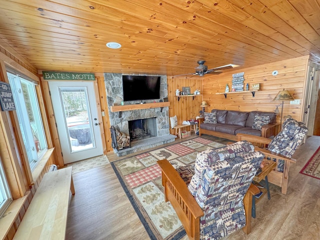 living area with a ceiling fan, wood finished floors, wooden walls, a stone fireplace, and wooden ceiling