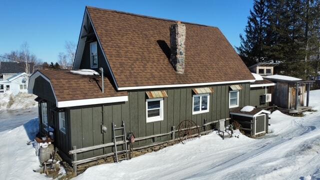 snow covered back of property featuring board and batten siding, roof with shingles, and a chimney