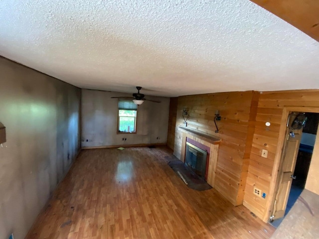 unfurnished living room with a brick fireplace, ceiling fan, wood walls, wood finished floors, and a textured ceiling