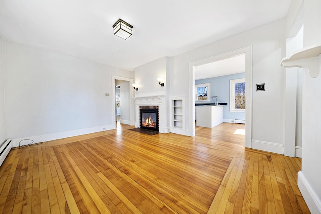 unfurnished living room featuring built in shelves, a baseboard heating unit, light wood-style floors, baseboards, and a brick fireplace