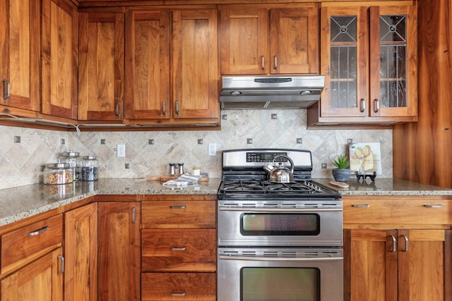 kitchen featuring tasteful backsplash, exhaust hood, brown cabinetry, range with two ovens, and light stone countertops