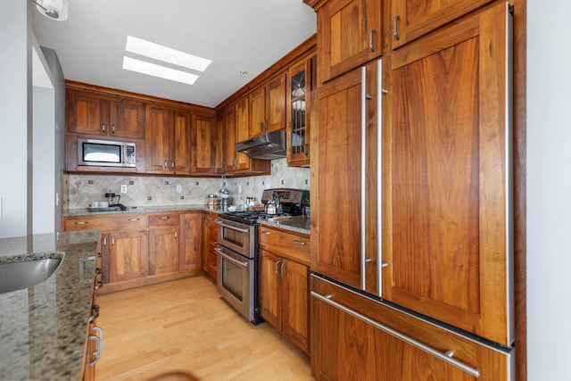 kitchen with under cabinet range hood, dark stone counters, appliances with stainless steel finishes, a skylight, and brown cabinetry