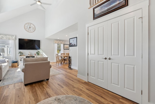 living area featuring a ceiling fan, light wood-style floors, baseboards, and high vaulted ceiling