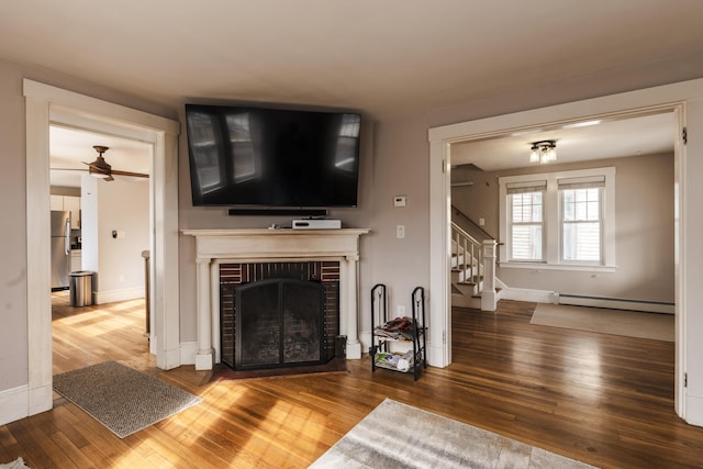 unfurnished living room with hardwood / wood-style flooring, stairway, a fireplace, a baseboard radiator, and ceiling fan