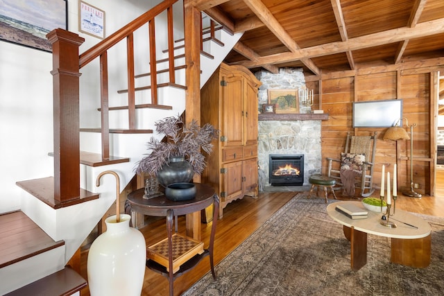 living area with wooden walls, beam ceiling, a stone fireplace, wooden ceiling, and wood finished floors