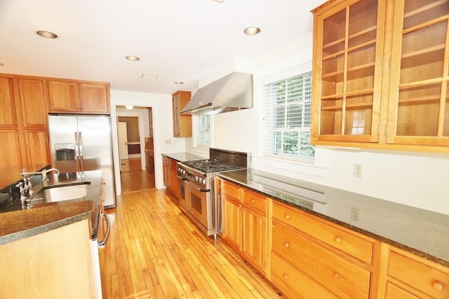 kitchen with dark stone countertops, a sink, appliances with stainless steel finishes, wall chimney exhaust hood, and light wood-type flooring