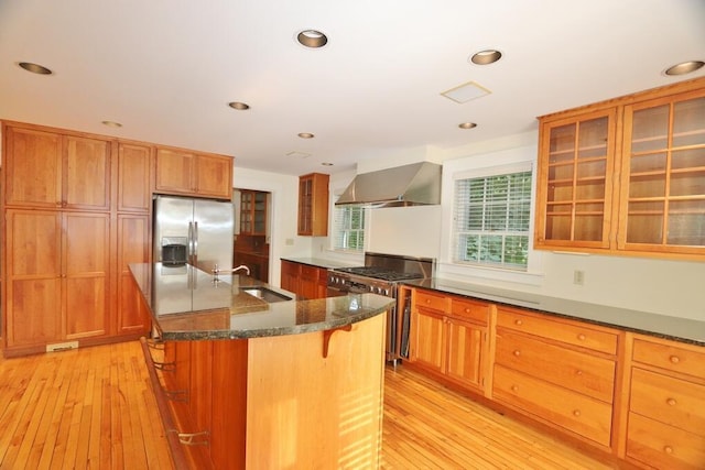kitchen with dark stone counters, an island with sink, stainless steel appliances, wall chimney exhaust hood, and a sink