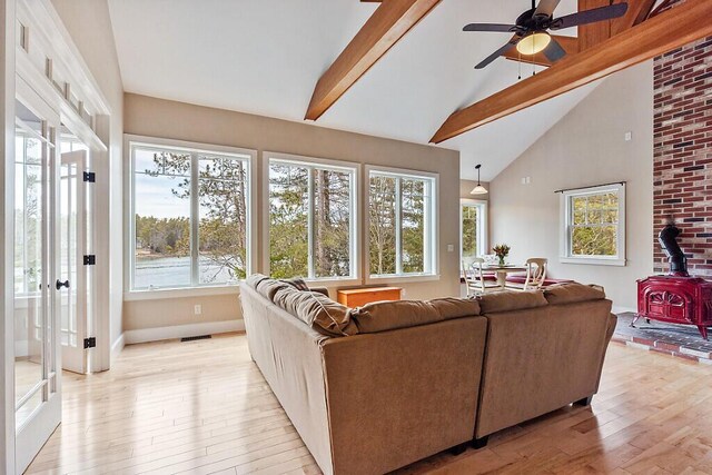 living room with ceiling fan, high vaulted ceiling, a wood stove, beamed ceiling, and light wood finished floors
