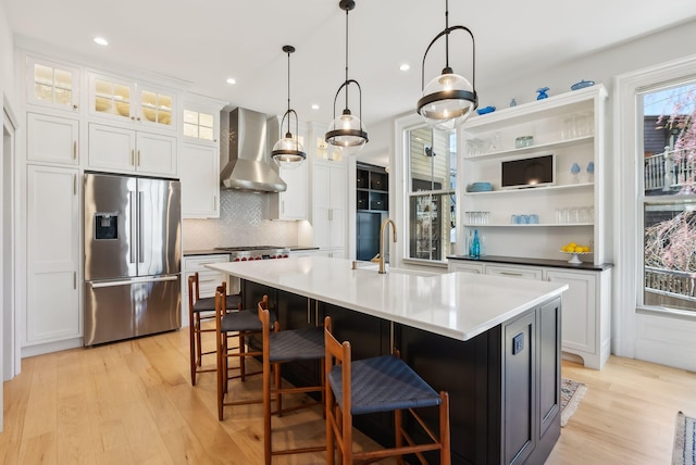 kitchen featuring light wood-style floors, wall chimney range hood, stainless steel refrigerator with ice dispenser, white cabinets, and a sink