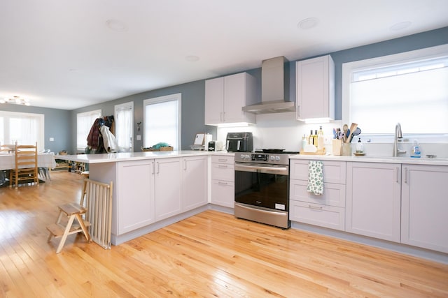 kitchen featuring stainless steel electric range, light wood finished floors, light countertops, wall chimney exhaust hood, and a peninsula