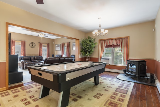 recreation room with a wood stove, a baseboard heating unit, wood finished floors, and ceiling fan with notable chandelier