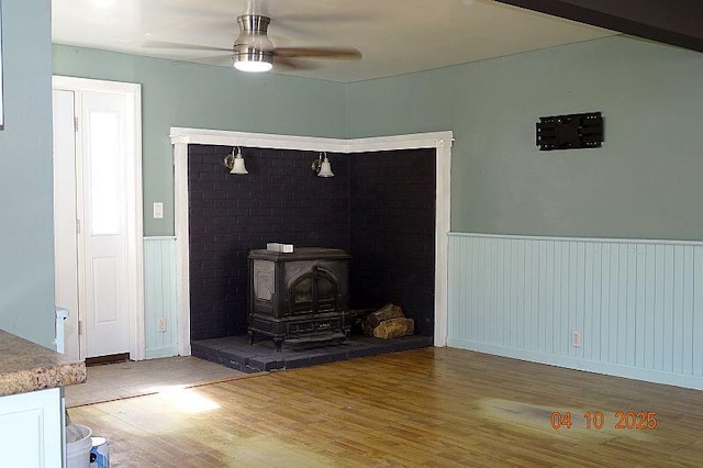 unfurnished living room with a wood stove, ceiling fan, wainscoting, and light wood-style floors