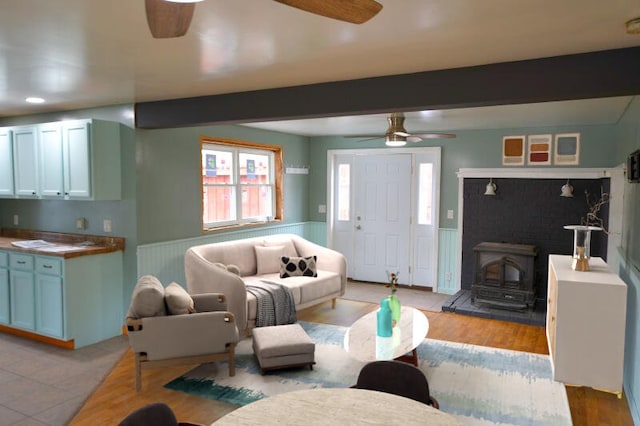 living room with wainscoting, light wood-style floors, beamed ceiling, a ceiling fan, and a wood stove