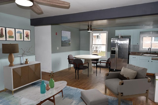 living area with beamed ceiling, a ceiling fan, and a wainscoted wall