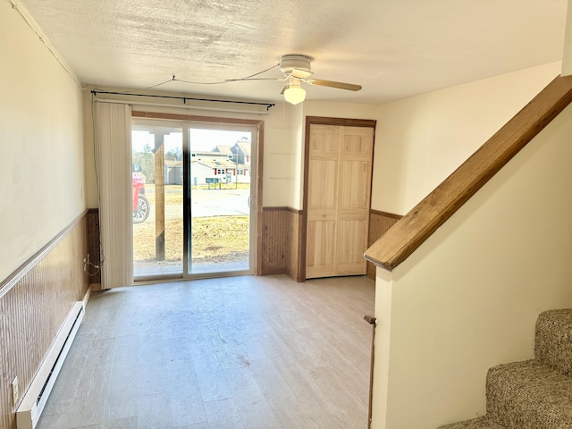 interior space with a wainscoted wall, light wood finished floors, stairway, a ceiling fan, and a baseboard heating unit