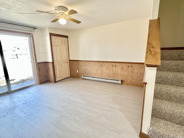 spare room with ceiling fan, stairway, baseboard heating, light wood-style floors, and wainscoting