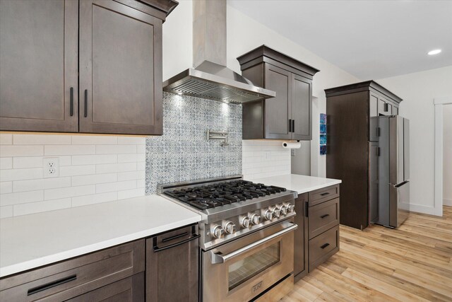 kitchen with stainless steel appliances, wall chimney exhaust hood, dark brown cabinets, light countertops, and light wood-style floors