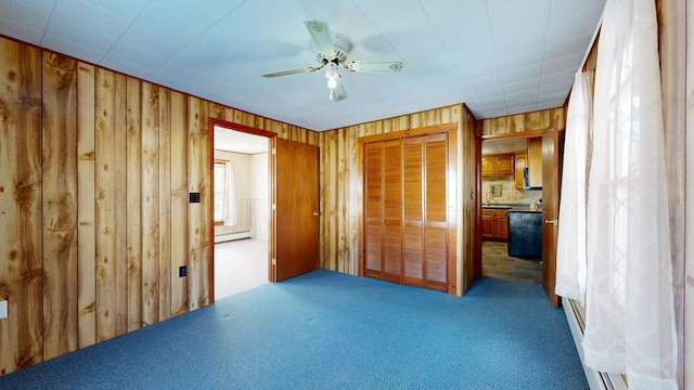 unfurnished bedroom featuring ceiling fan, baseboard heating, ensuite bath, wood walls, and carpet flooring
